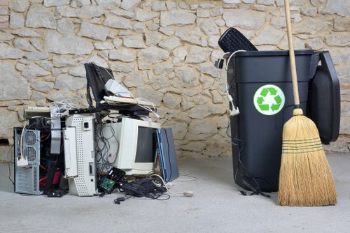 In the foreground, a pile of discarded electronic waste, including old computer towers, keyboards, and cables, appears disorganized and partially stacked, with some components spilling onto the concrete surface. The electronic waste displays various textures, with metal casings, plastic shells, and tangled wiring, predominantly in shades of gray, black, and metallic silver. To the right, a black wheelie bin with a green recycling symbol is positioned against a beige stone wall, with a wooden broom leaning against it, its bristles resting on the ground. The environment suggests an outdoor area, possibly a driveway or back entrance, where rubbish collection or removal services are carried out. The scene is lit with natural daylight, casting soft shadows that highlight the textures of the waste materials and the stone wall background. This visual is relevant for a rubbish removal service such as Commercial Waste Farnborough, emphasizing the handling of electronic waste and general rubbish disposal in the Farnborough area.