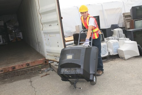 A woman wearing a yellow safety helmet, high-visibility vest with red and yellow stripes, and work gloves is standing outdoors on a concrete surface near an open storage container or warehouse. She is holding and positioning a black electronic device, which appears to be a portable or industrial piece of equipment, with the front panel featuring vents, buttons, and a display. Behind her, there are stacks of various waste materials, including old computer monitors, printers, and other electronic equipment, arranged along the edge of the storage area. The background shows a white curved structure, possibly a temporary shelter or tent, and the scene suggests an environment focused on waste management or recycling. The overall setting indicates a commercial waste removal operation, consistent with services provided by companies like Commercial Waste Farnborough, operating in the Farnborough area postcode. The lighting appears natural, highlighting the functional and professional nature of the activity related to waste handling and electronics disposal.