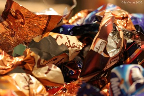 A close-up of crumpled metallic foil wrappers and plastic packaging, predominantly in shades of copper, silver, and dark blue, with some printed text and colorful branding visible. The wrappers are layered and tangled, creating a textured, glossy surface reflecting light. The background appears blurred, focusing entirely on the waste materials, which are typically associated with discarded confectionery or snack wrappers. The image emphasizes the need for rubbish removal services like those provided by Commercial Waste Farnborough, especially for handling small packaged waste items in commercial or household settings within the Farnborough postcodes. The cluttered appearance underscores the importance of proper disposal and waste collection in local waste management efforts.